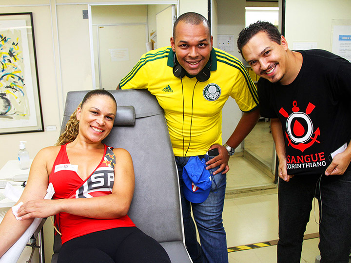 Dois homens e uma mulher vestindo camisas de times de futebol sorriem enquanto a mulher faz uma doação de sangue